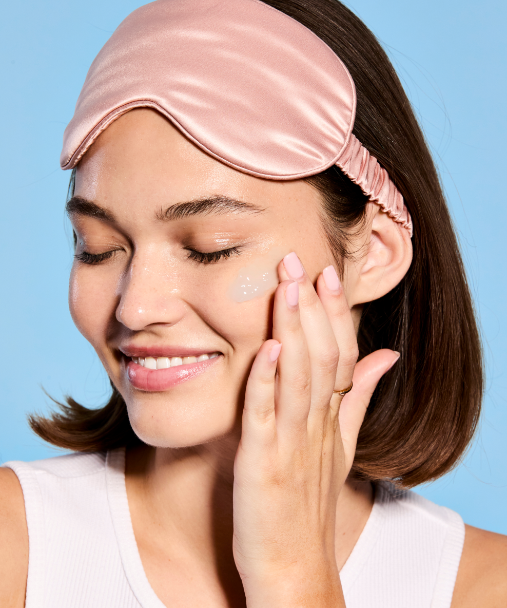 A smiling model wearing an eye mask on her forehead rubs some belif Aqua Bomb Sleeping Mask onto her cheek