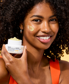 A model smiles while holding an open jar of belif Aqua Bomb Brightening Vitamin C Cream with some cream on her cheek