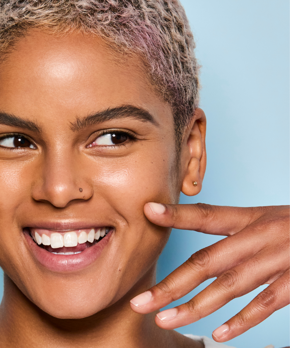 A model with glowing skin smiles as she lightly touches her cheek with her finger