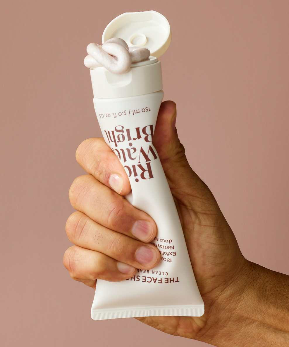 A hand squeezing a tube of The Face Shop Rice Water Bright Rice Bran Gentle Exfoliating Cleanser, highlighting its vegan formula and skin-brightening benefits